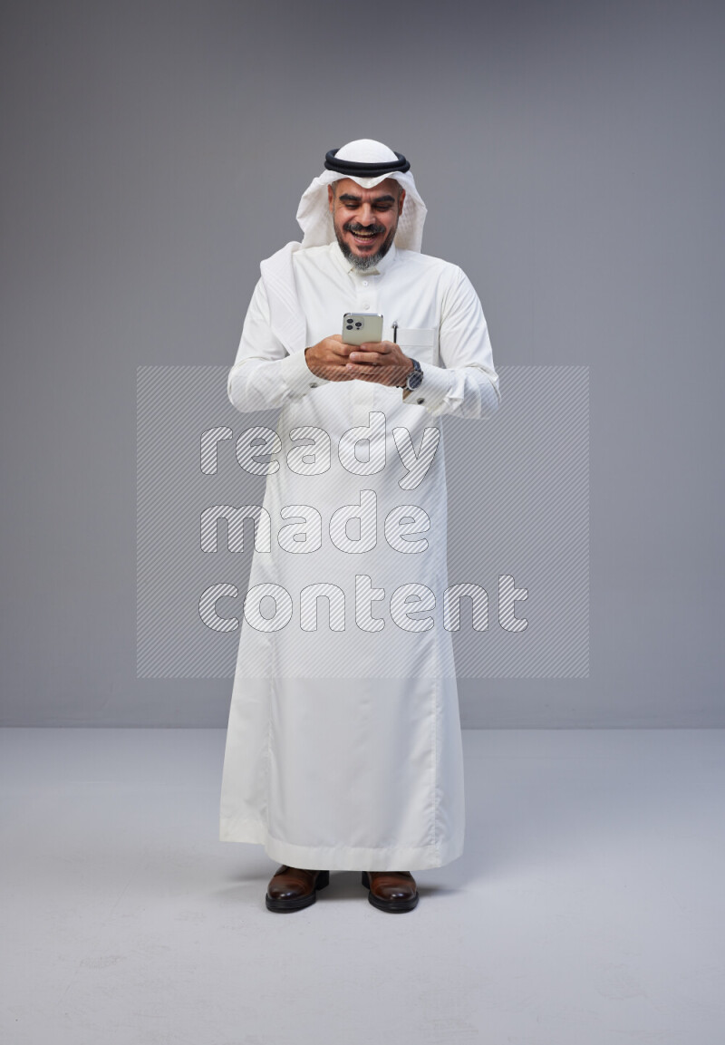 Saudi man Wearing Thob and white Shomag standing texting on phone on Gray background