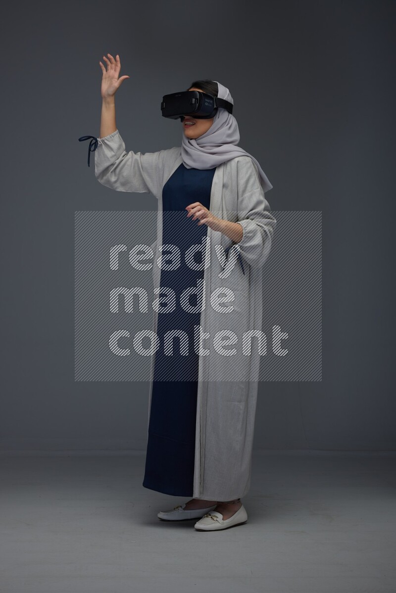A Saudi woman wearing a light gray Abaya and head scarf standing and wearing VR eye level on a grey background