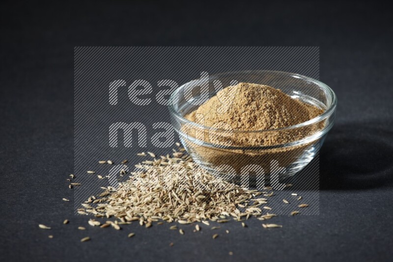 A glass bowl full of cumin powder with cumin seeds beside it on black flooring