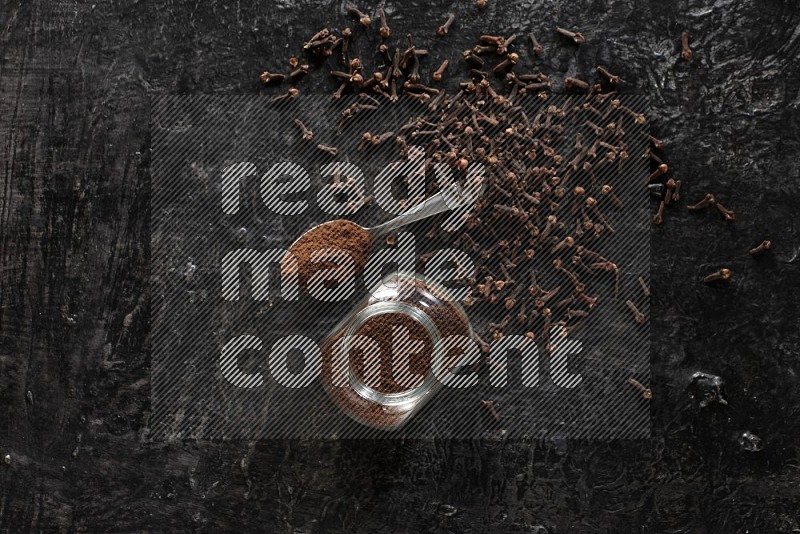 A glass spice jar and a metal spoon full of cloves powder with cloves spread on textured black flooring