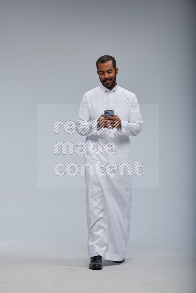 Saudi man wearing Thob standing texting on phone on Gray background
