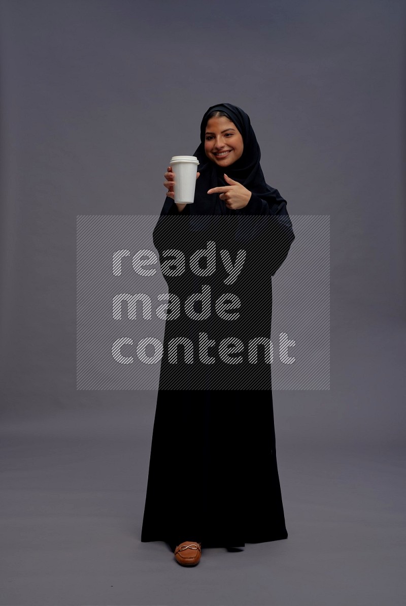 Saudi woman wearing Abaya standing holding paper cup on gray background