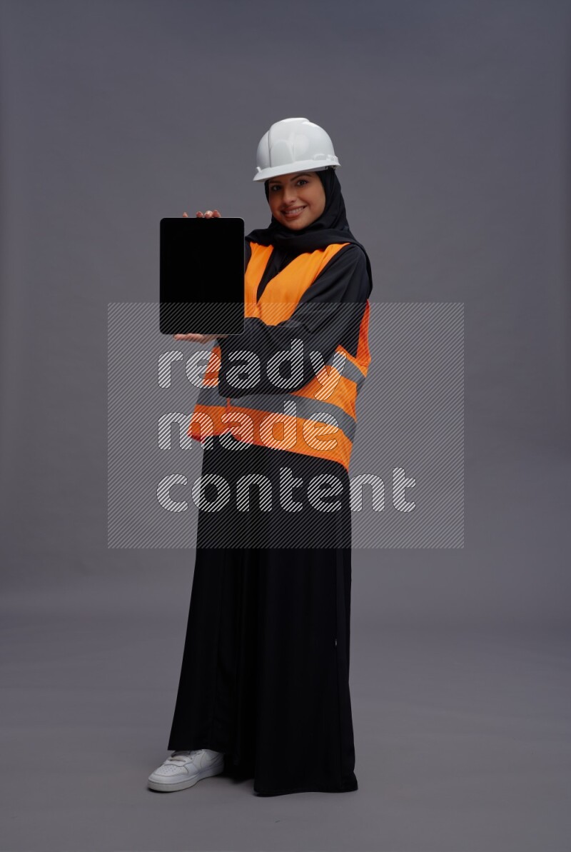 Saudi woman wearing Abaya with engineer vest standing showing tablet to camera on gray background