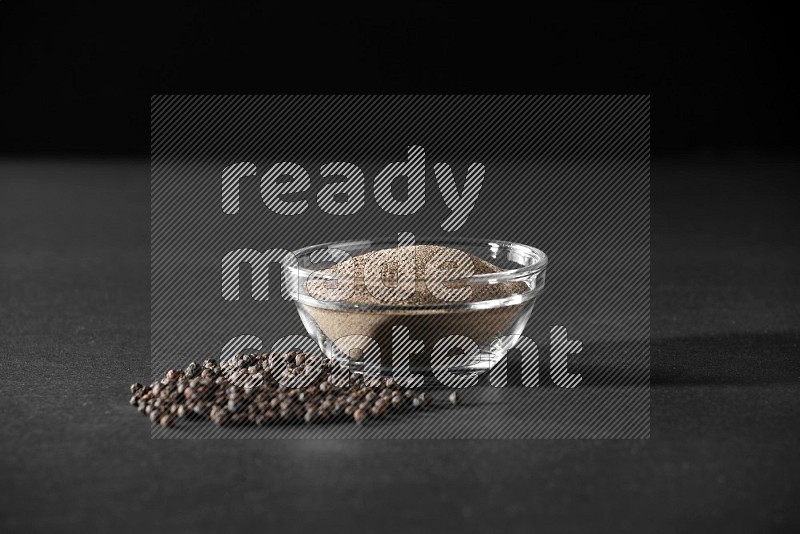 A glass bowl full of powder black pepper powder and black pepper beads on the floor on black flooring