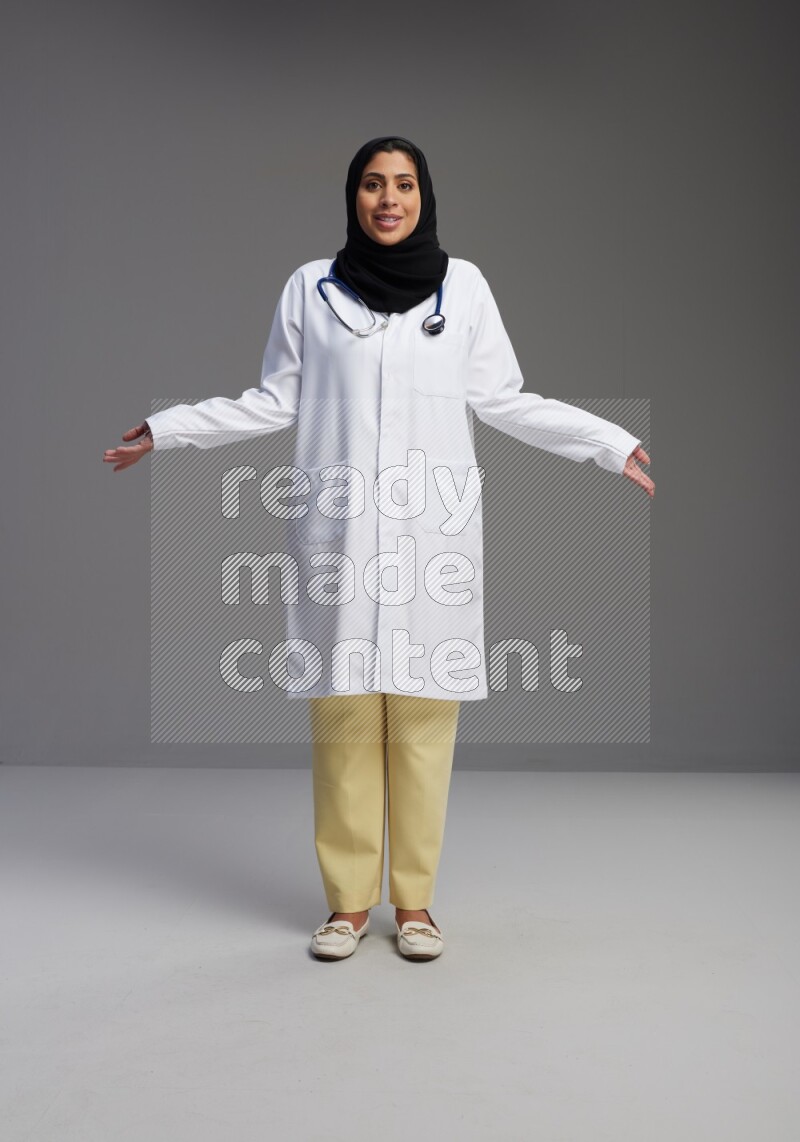 Saudi woman wearing lab coat with stethoscope standing interacting with the camera on Gray background