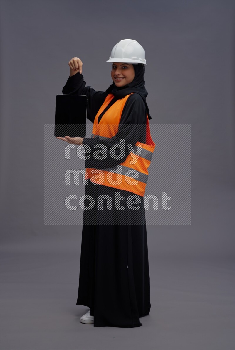 Saudi woman wearing Abaya with engineer vest standing showing tablet to camera on gray background
