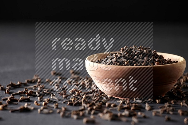 A wooden bowl full of cloves with spreaded whole cloves on a black flooring