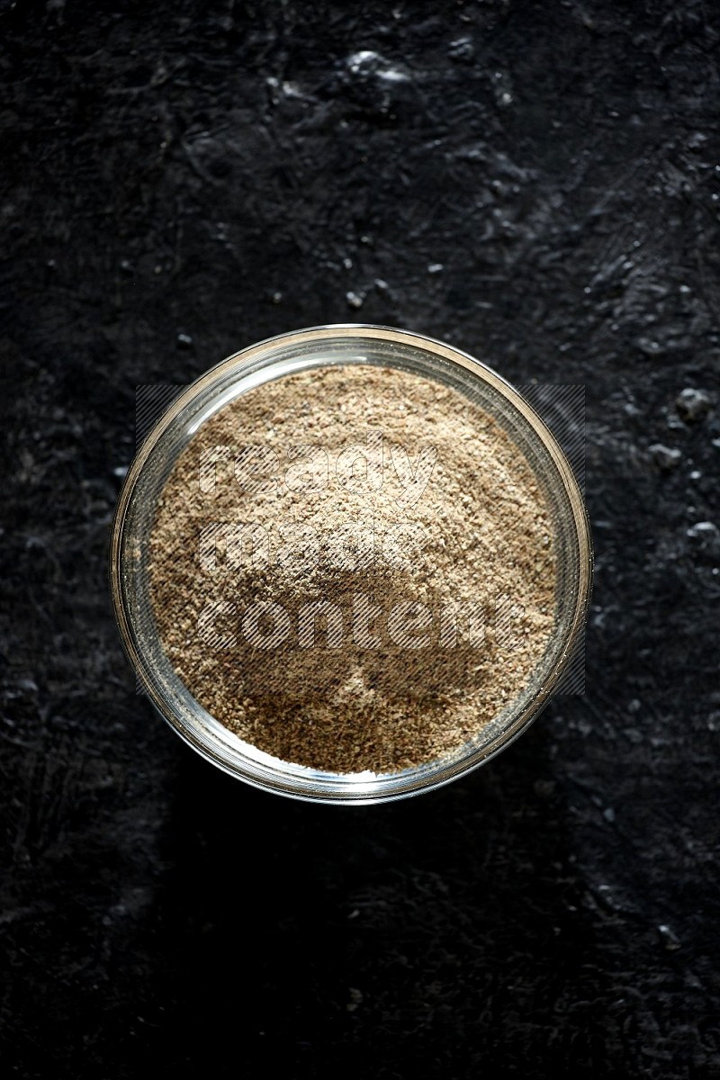 A glass bowl full of cardamom powder on textured black flooring