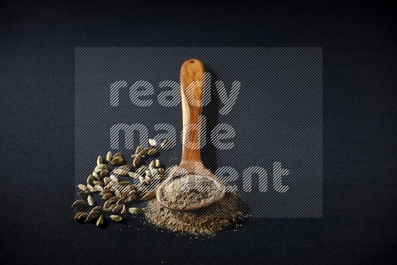 A wooden ladle full of cardamom powder and cardamom seeds beside it on black flooring