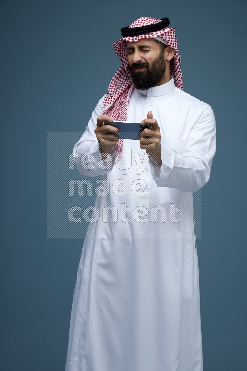 A man Playing a Game on his phone in a blue background wearing Saudi Thob with Shomag