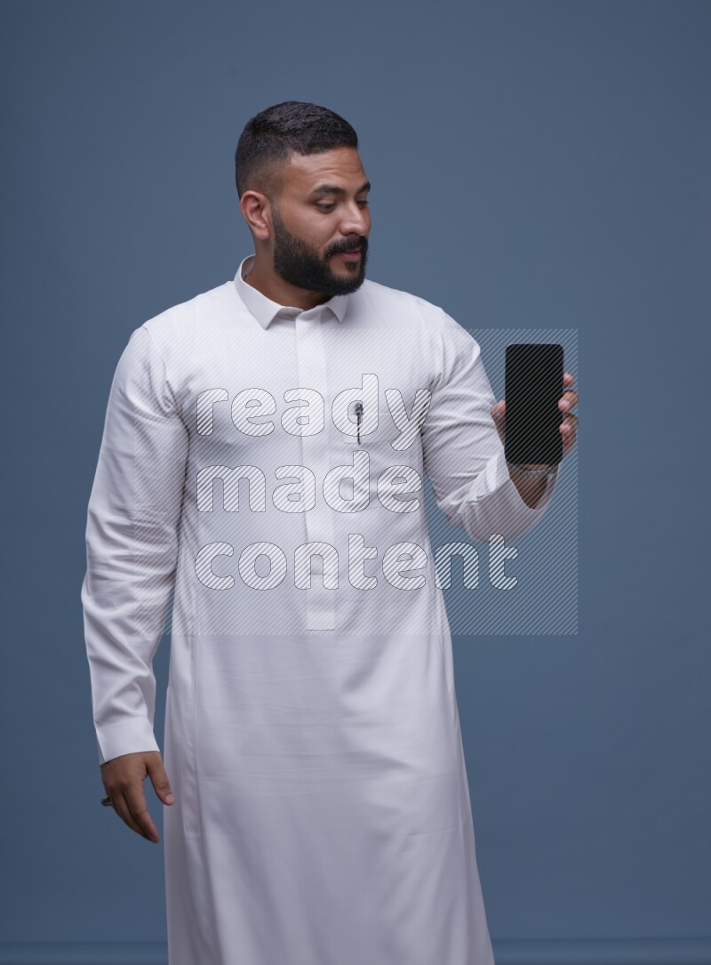 A man Showing his phone in Blue Background wearing Saudi Thob