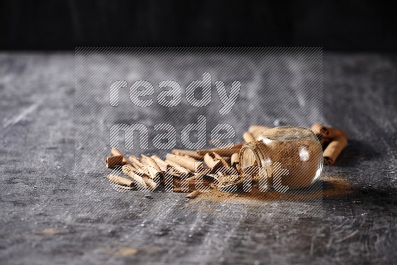 Herbal glass jar full cinnamon powder flipped and a metal spoon full of powder surrounded by cinnamon sticks on textured black background in different angles