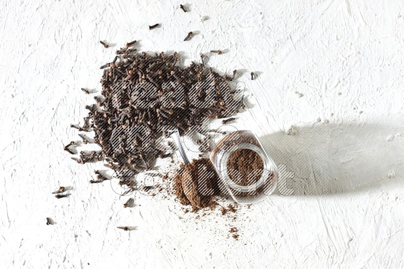 A glass spice jar and a metal spoon full of cloves powder and cloves spread on textured white flooring