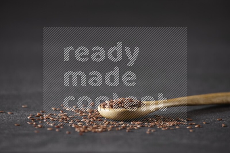 A wooden spoon full of flaxseeds and seeds spread beside it on a black flooring