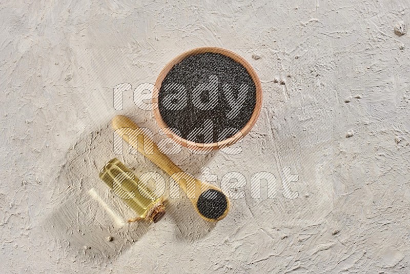 A wooden bowl and spoon full of black seeds with a bottle of black seeds oil on a textured white flooring