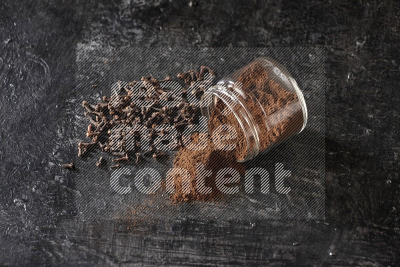 A flipped glass jar full of cloves powder with cloves spread on a textured black flooring