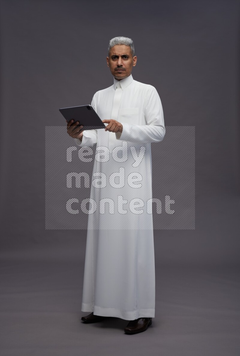 Saudi man wearing thob standing working on tablet on gray background