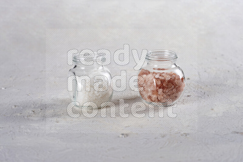 2 glass jars one is filled with coarse himalayan salt and the other with coarse sea salt on white background