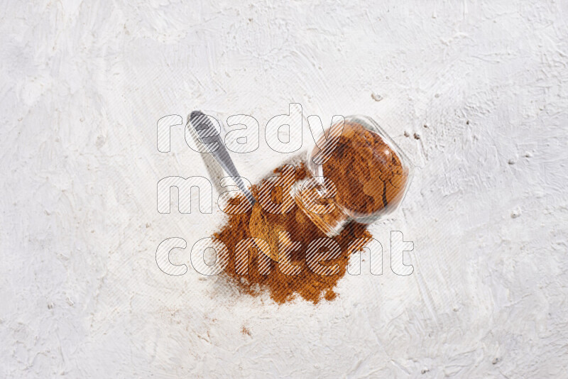 A glass jar full of ground paprika powder flipped with some spilling powder on white background