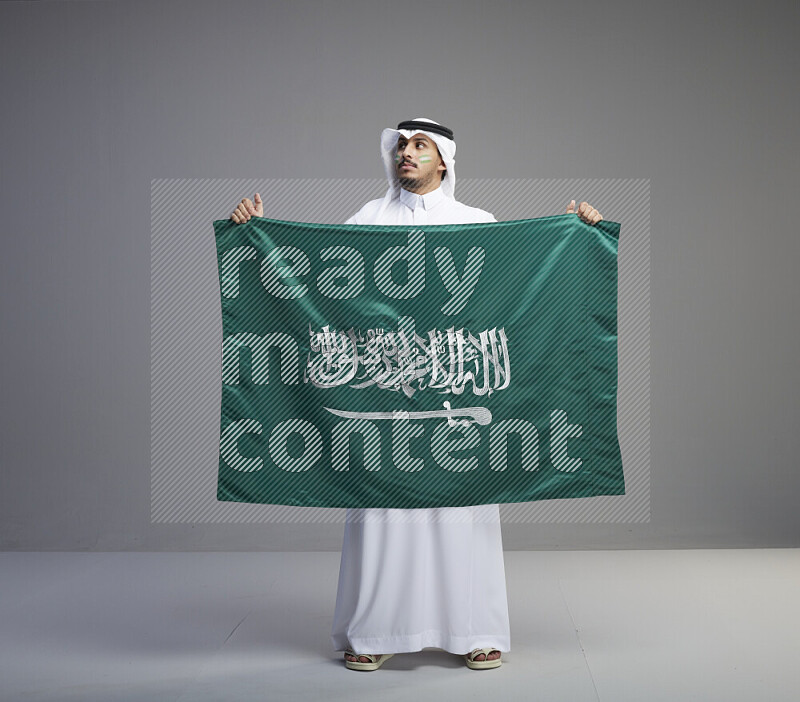 A Saudi man standing wearing thob and white shomag with face painting holding big Saudi flag on gray background