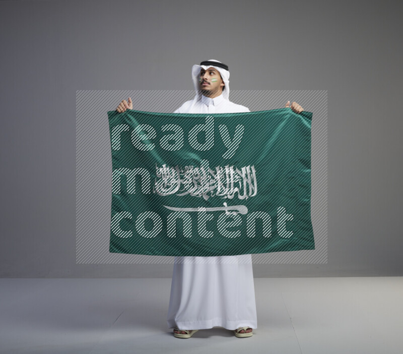 A Saudi man standing wearing thob and white shomag with face painting holding big Saudi flag on gray background