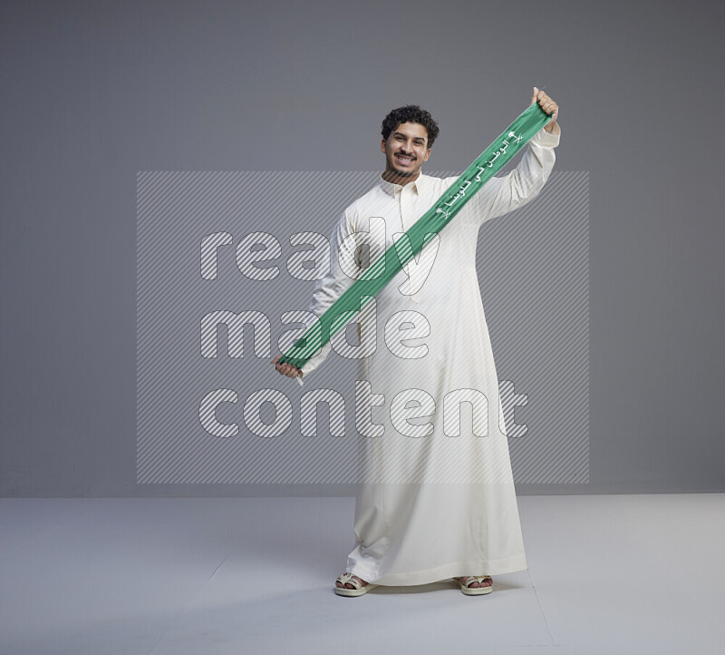 A Saudi man standing wearing thob holding Saudi flag scarf on gray background