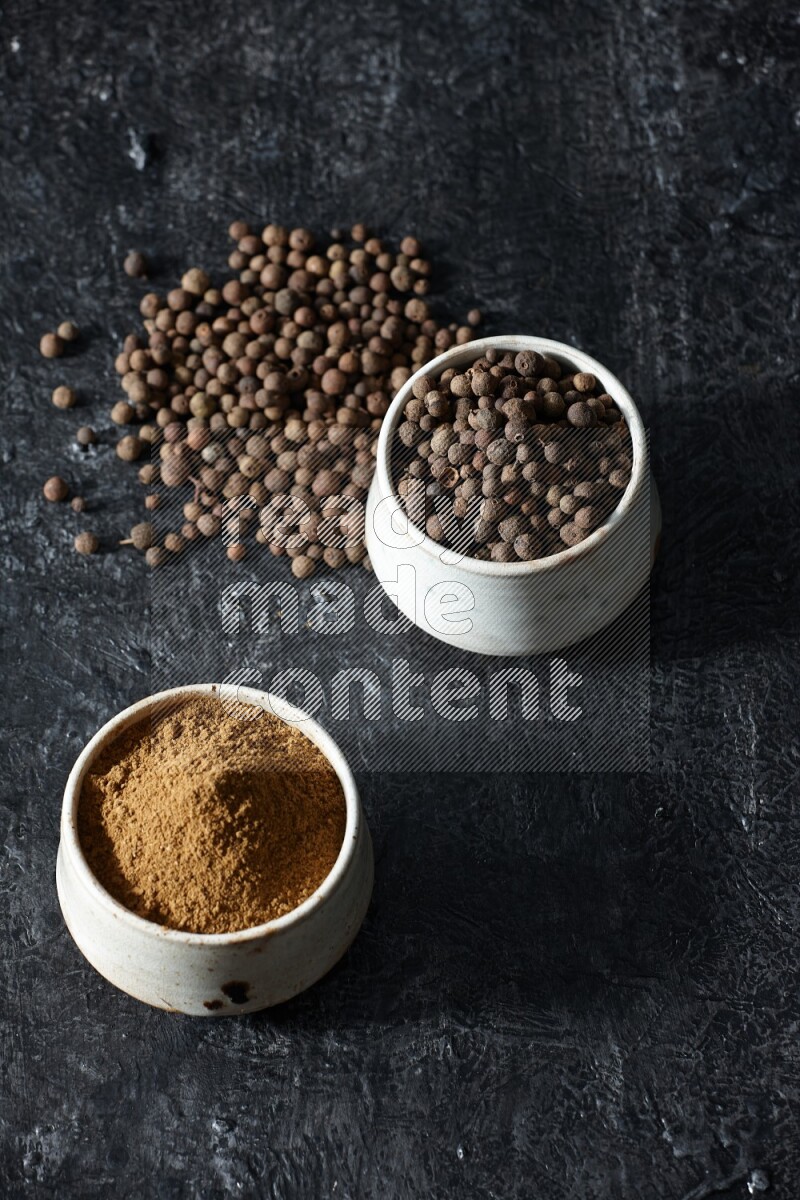 2 beige bowls, one full of allspice powder and the other full of whole balls and both spread on a textured black flooring
