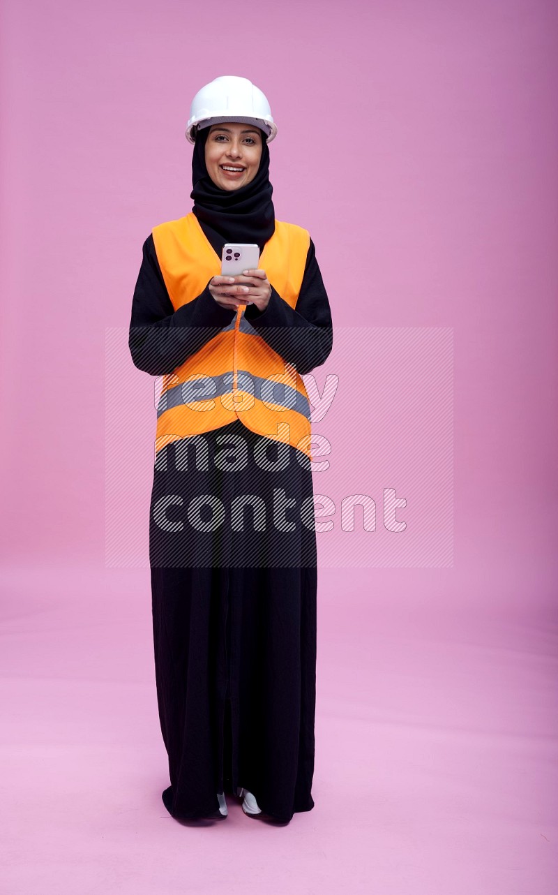 Saudi woman wearing Abaya with engineer vest and helmet standing texting on phone on pink background