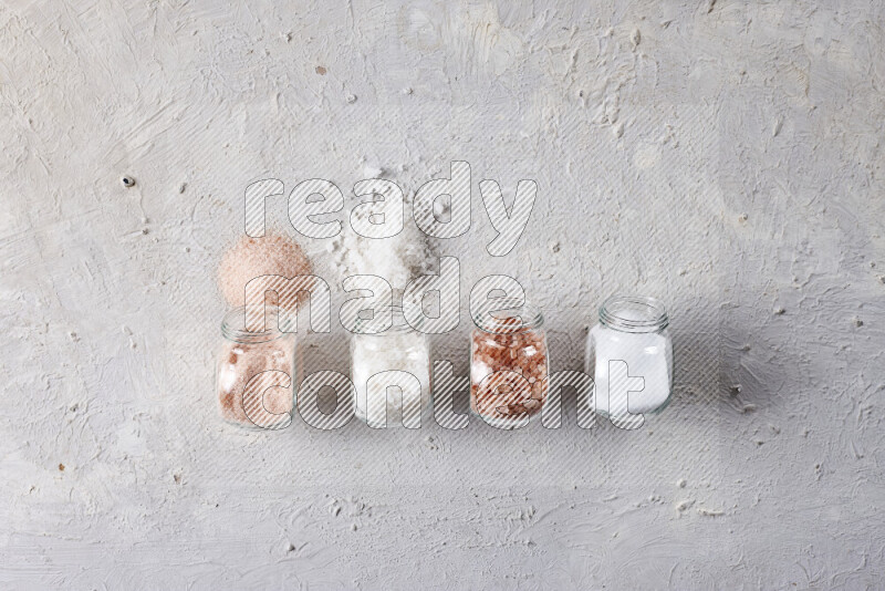 4 glass jars filled with table salt, coarse sea salt, fine himalayan salt and coarse himalayan salt on white background