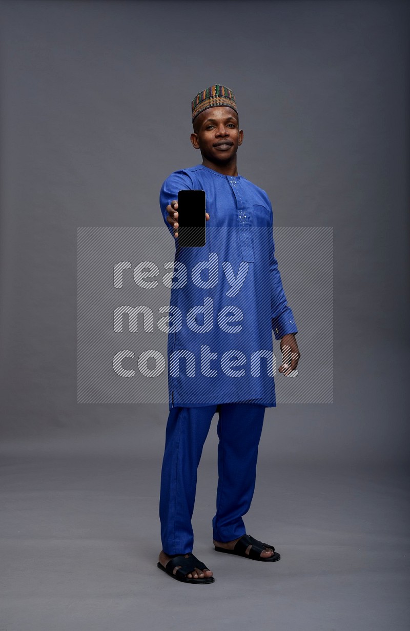 Man wearing Nigerian outfit standing showing phone to camera on gray background