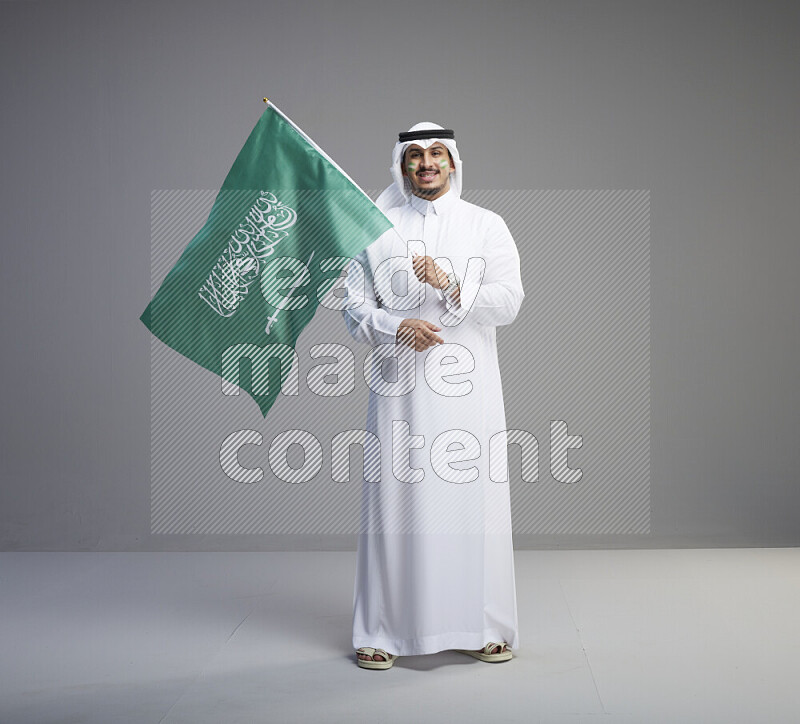 A Saudi man standing wearing thob and white shomag with face painting raising big Saudi flag on gray background