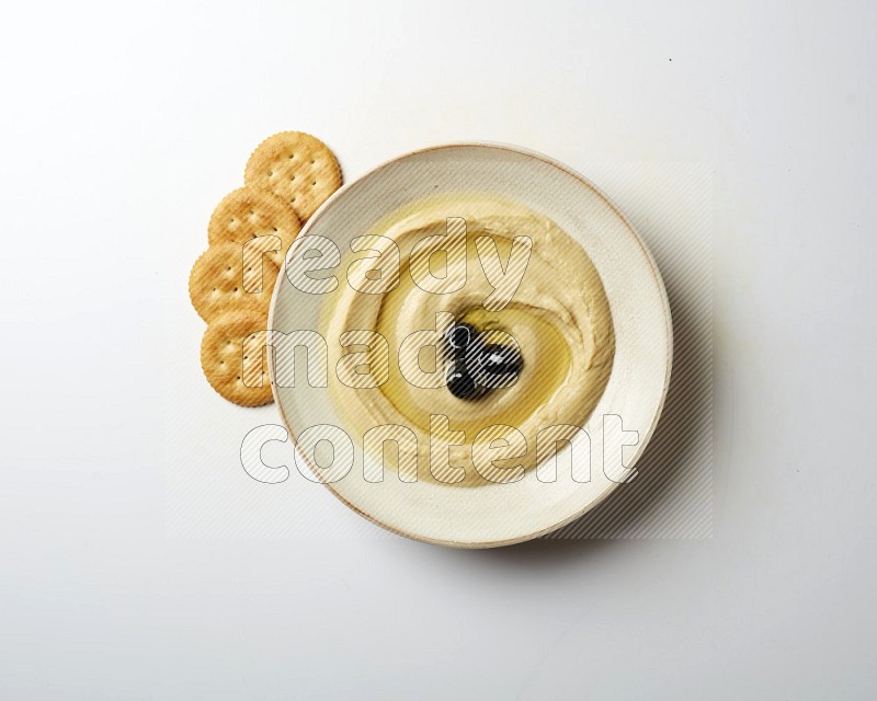 Hummus in a pottry plate garnished with black olives on a white background