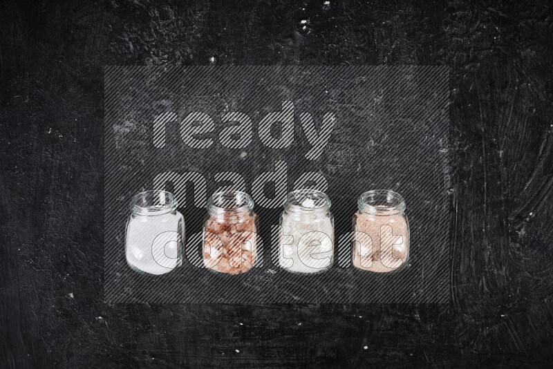 4 glass jars filled with table salt, coarse sea salt, fine himalayan salt and coarse himalayan salt on black background