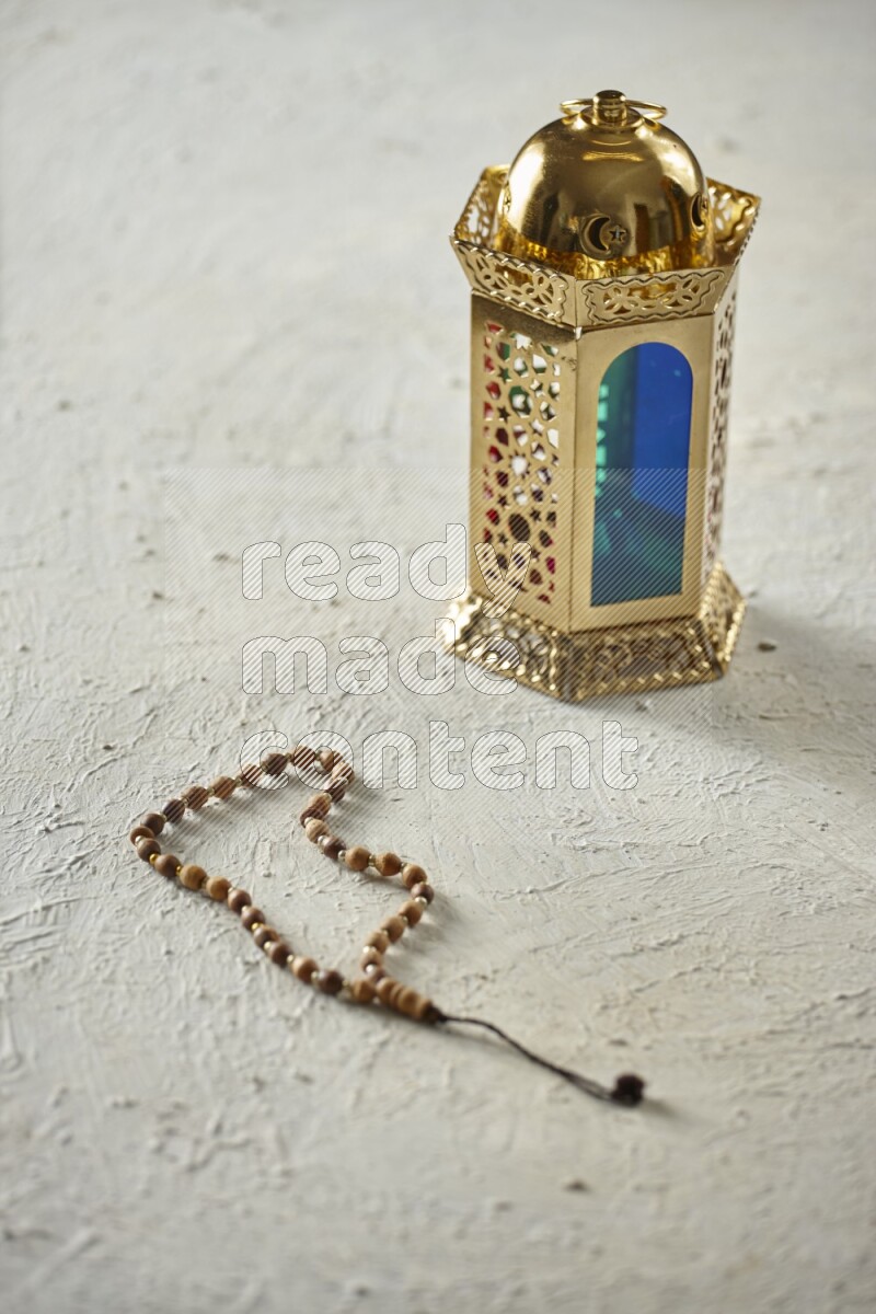 A golden lantern with different drinks, dates, nuts, prayer beads and quran on textured white background