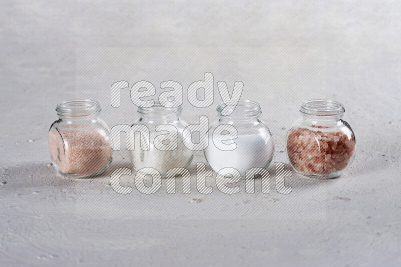 4 glass jars filled with table salt, coarse sea salt, fine himalayan salt and coarse himalayan salt on white background