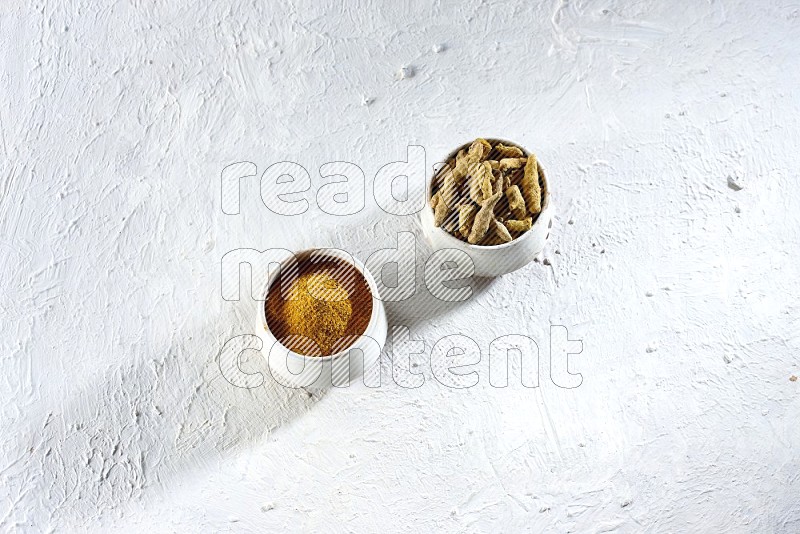 2 beige pottery bowl full of turmeric powder and dried turmeric whole fingers on textured white flooring
