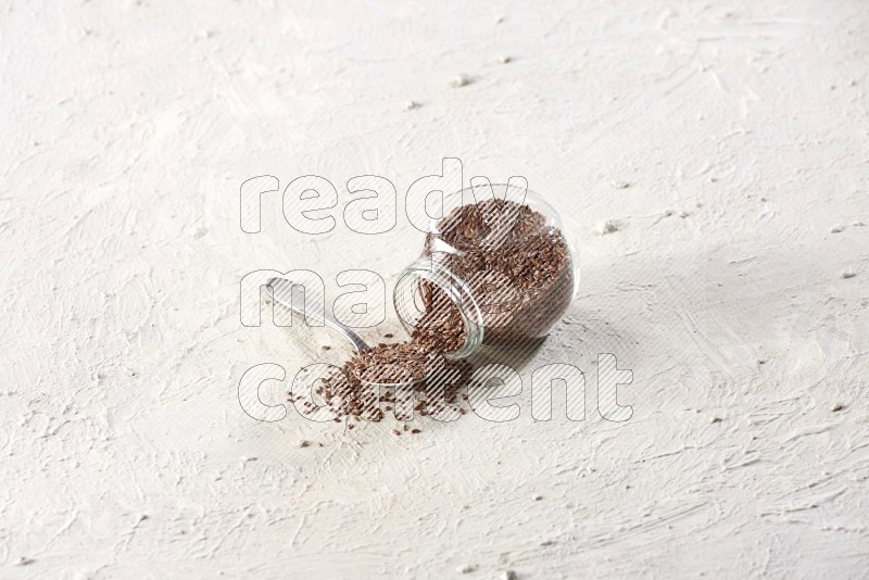 A glass spice jar full of flax flipped and a metal spoon full of the seeds on a textured white flooring in different angles