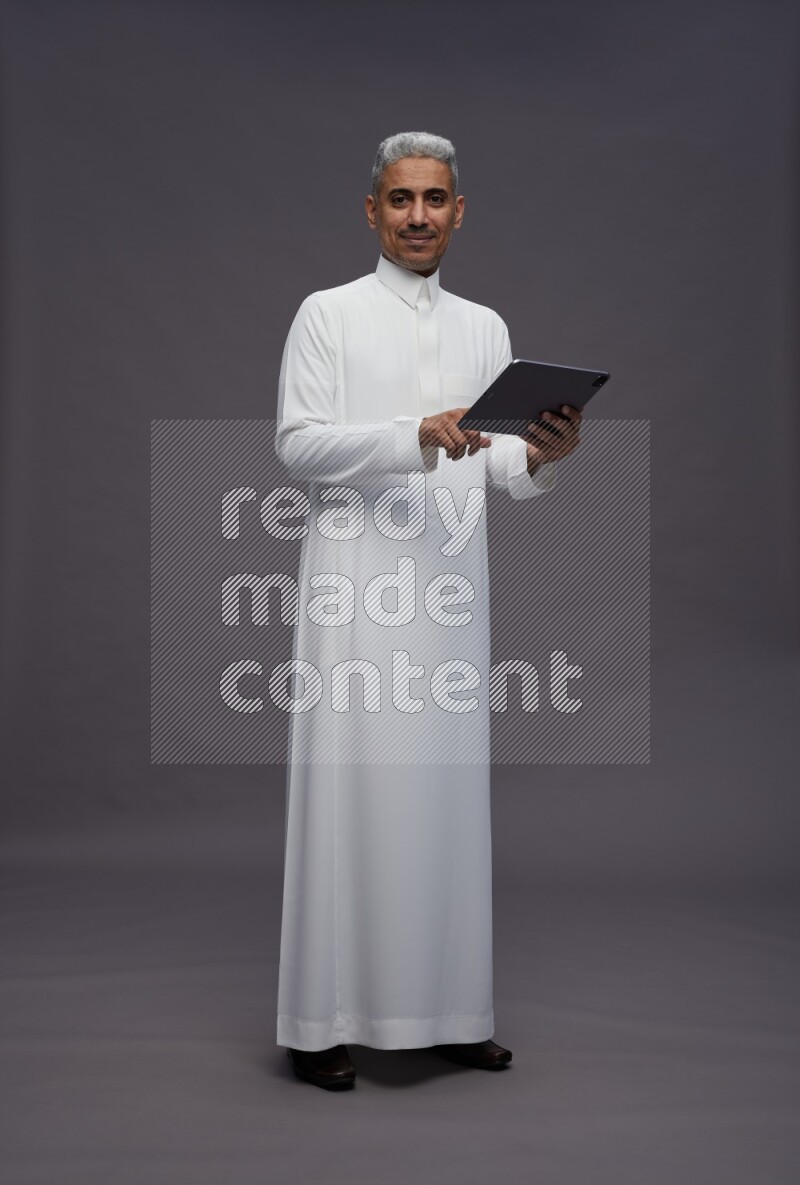 Saudi man wearing thob standing working on tablet on gray background