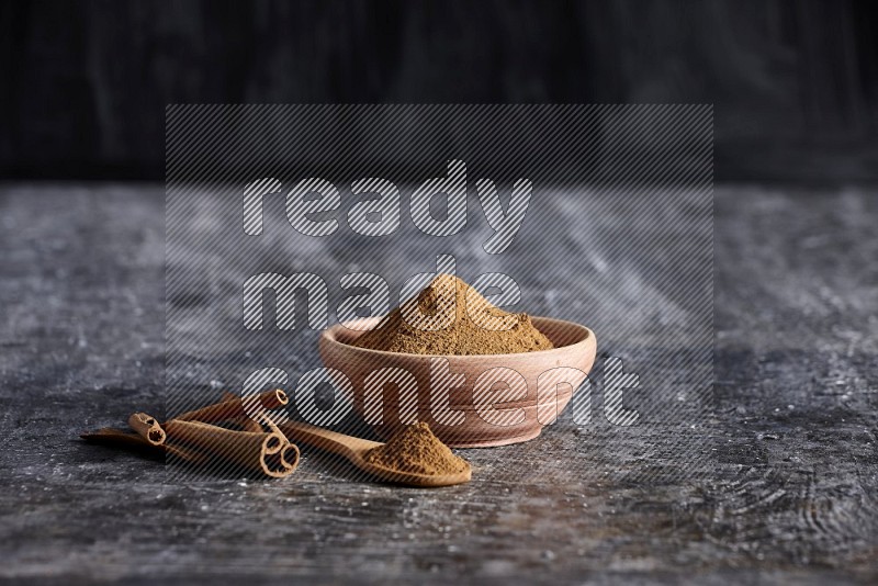 wooden bowl full of cinnamon powder and a wooden spoon full of it with cinnamon sticks on a textured black background