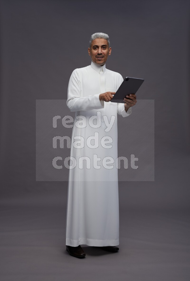 Saudi man wearing thob standing working on tablet on gray background