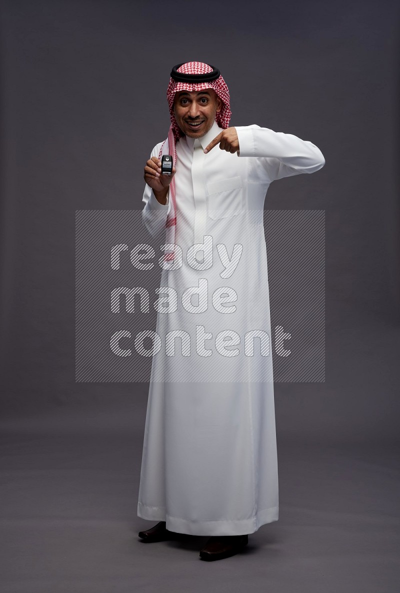 Saudi man wearing thob and shomag standing holding key car on gray background
