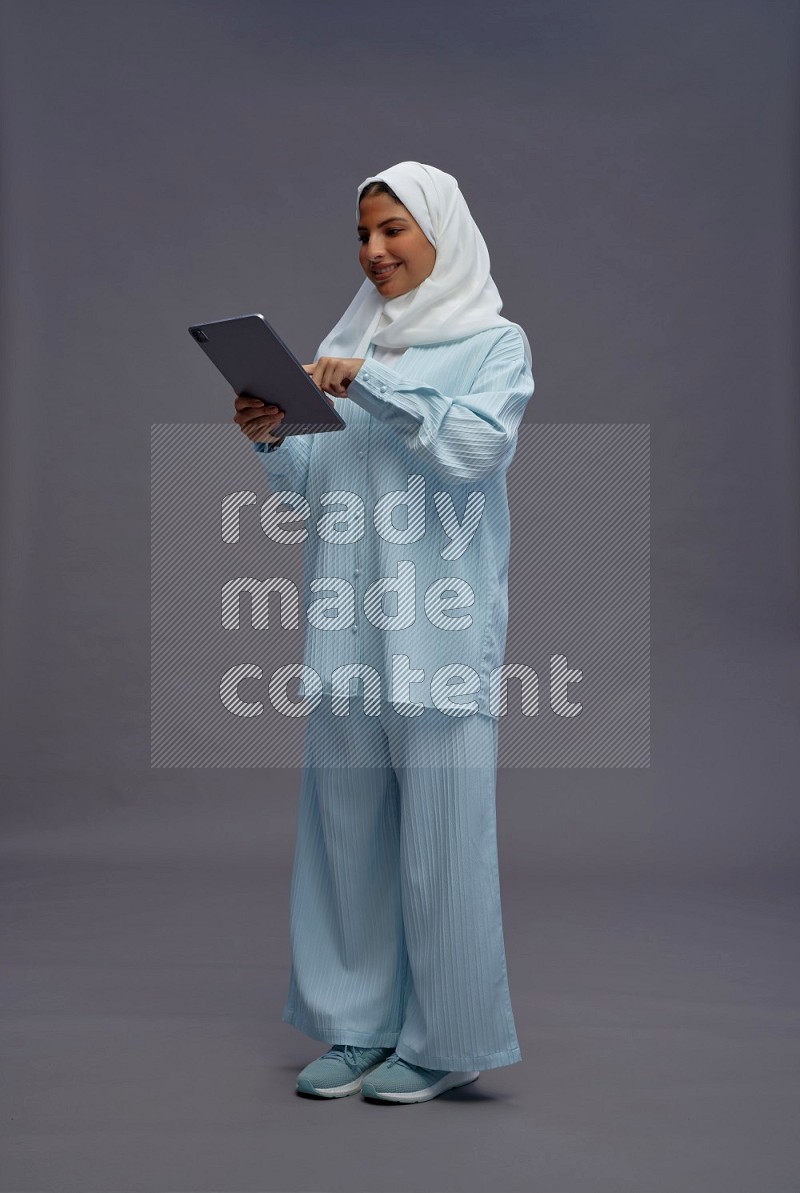 Saudi woman wearing hijab clothes standing working on tablet on gray background