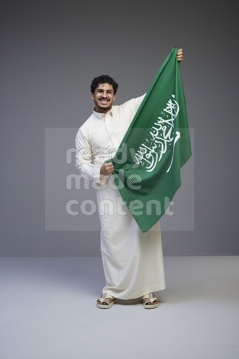 A saudi man standing wearing thob holding big saudi flag on gray background