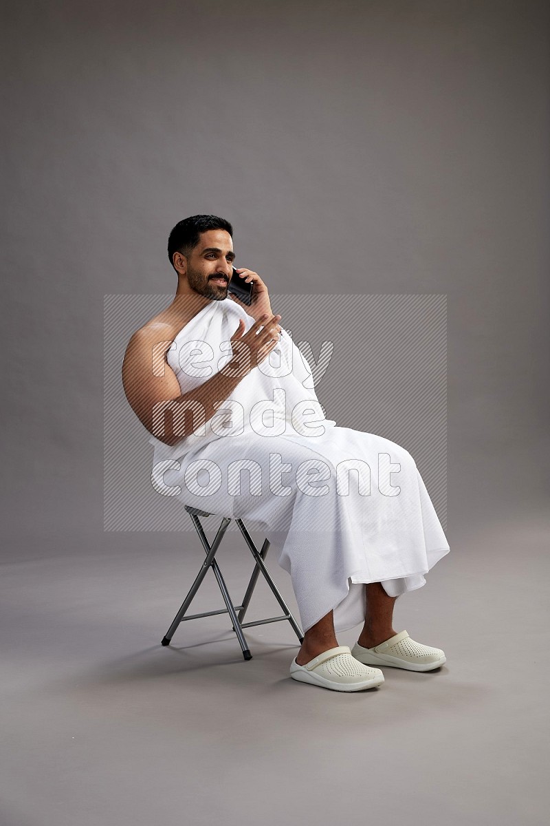 A man wearing Ehram sitting on chair talking on the phone on gray background