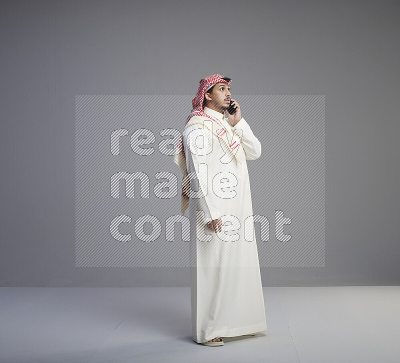 A Saudi man standing wearing thob and red shomag talking on phone on gray background