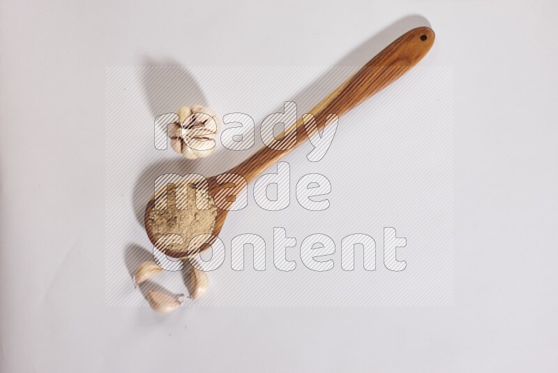 A wooden ladle full of garlic powder with a whole garlic bulb and some cloves beside it on a white flooring