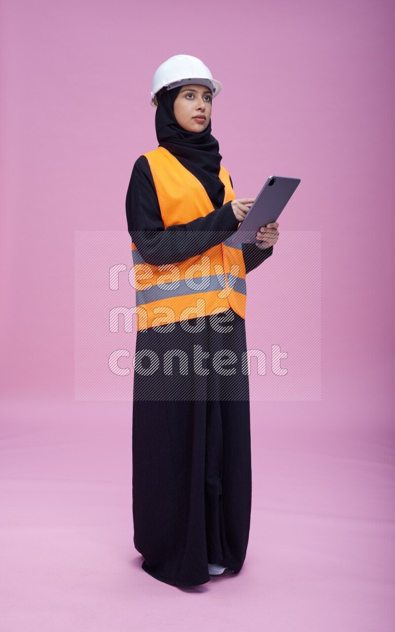 Saudi woman wearing Abaya with engineer vest and helmet standing working on tablet on pink background