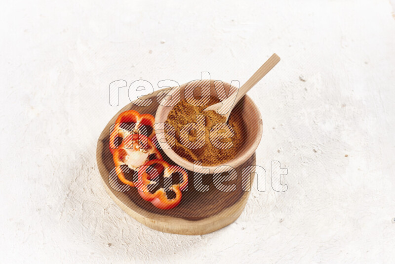 A wooden bowl full of ground paprika powder and sliced red bell pepper beside it all on a wooden tray on white background