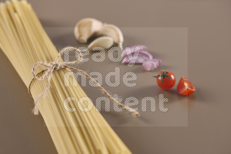 Raw pasta with different ingredients such as cherry tomatoes, garlic, onions, red chilis, black pepper, white pepper, bay laurel leaves, rosemary and cardamom on beige background