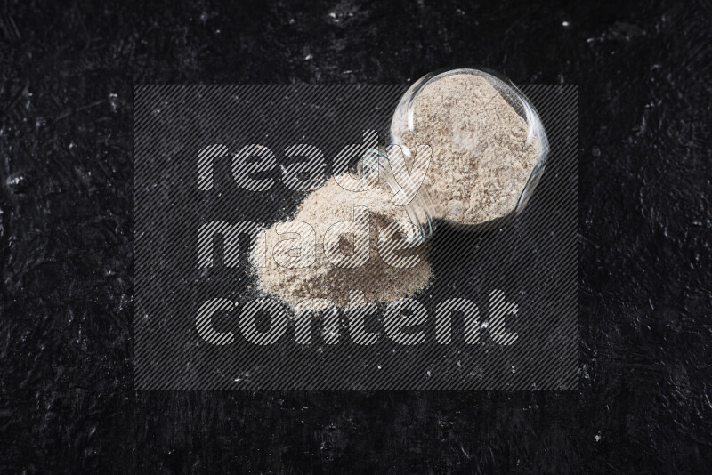 A glass jar full of onion powder flipped with some spilling powder on black background